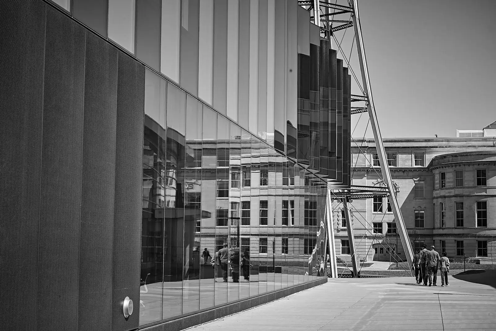 Black and white view of a glass building reflecting a stone building, with people walking across the plaza