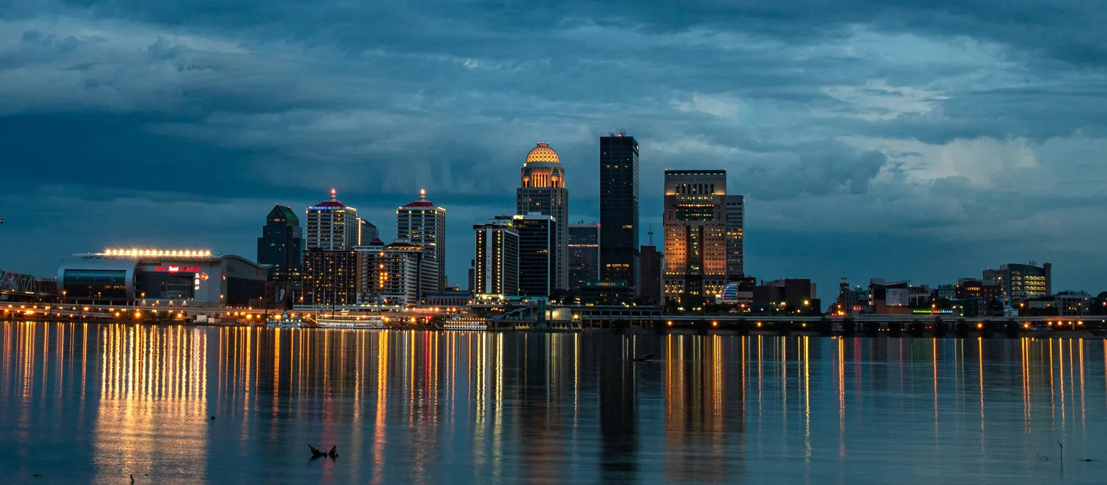 Downtown Louisville skyline at dusk across the Ohio River, with lit towers and the waterfront arena reflected in the water