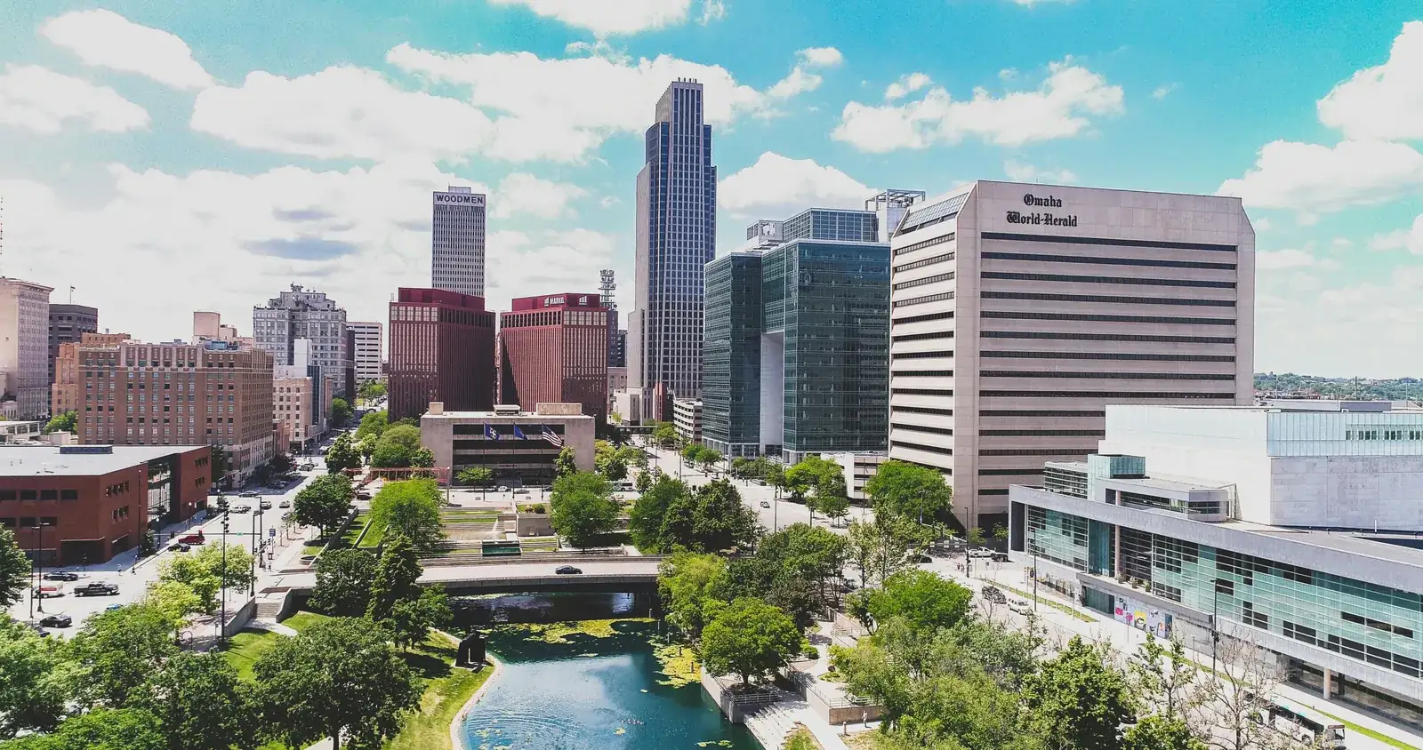 Aerial view of downtown Omaha on a sunny day, with office towers above a tree-lined park and lagoon