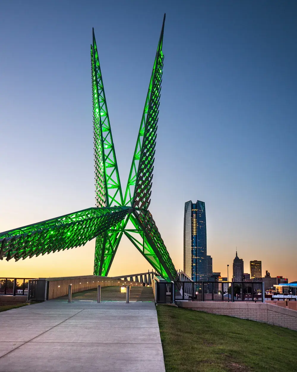 Green-lit Skydance Bridge sculpture at dusk with the downtown Oklahoma City skyline behind it