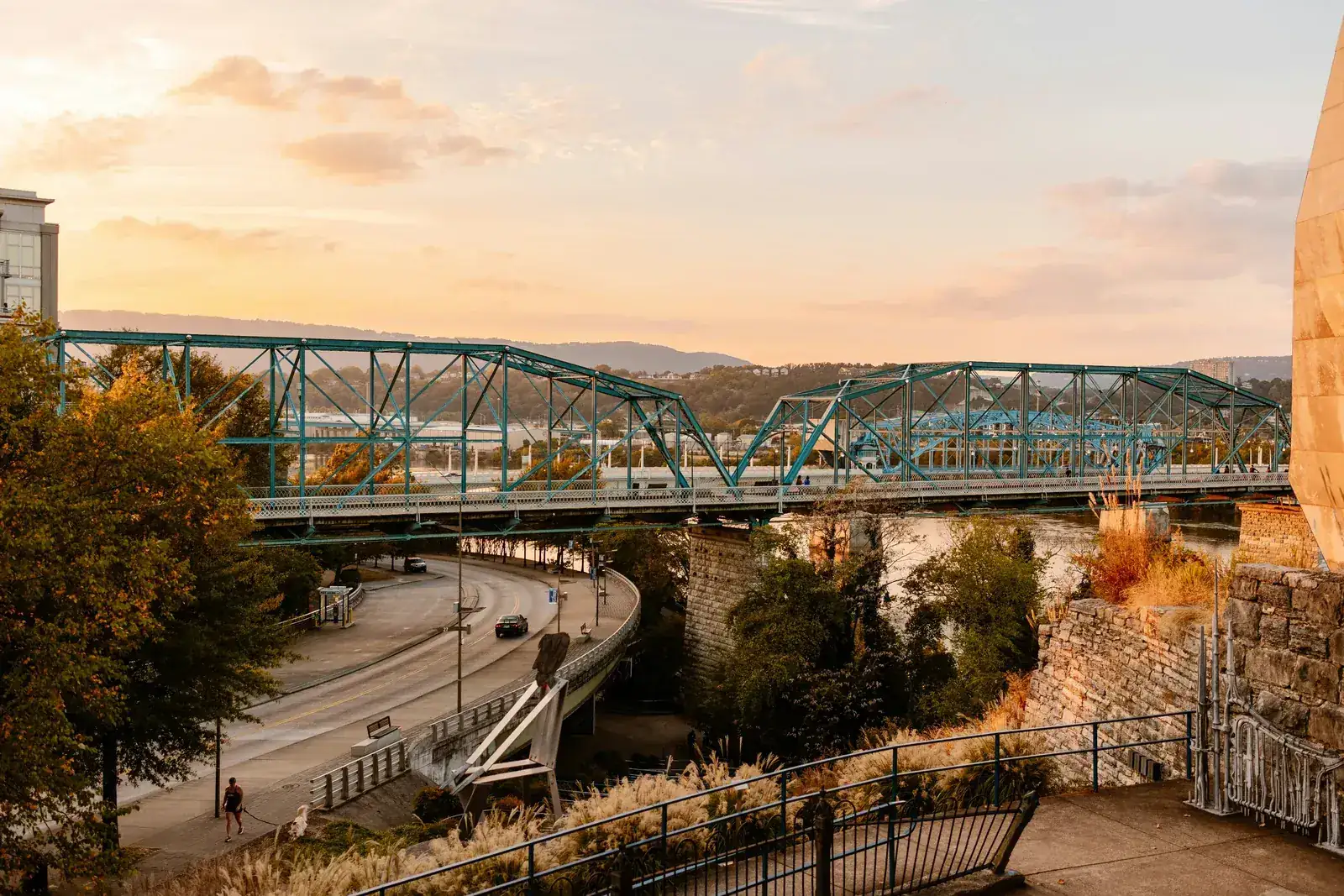 Teal steel truss bridge crossing a river at sunset, above a curving riverside road and autumn trees