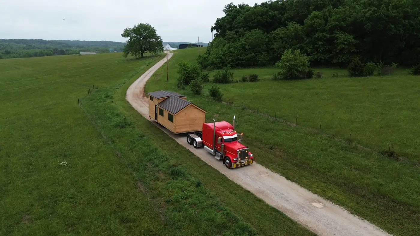 Aerial view of a red semi truck hauling a cabin down a long drive through open pasture