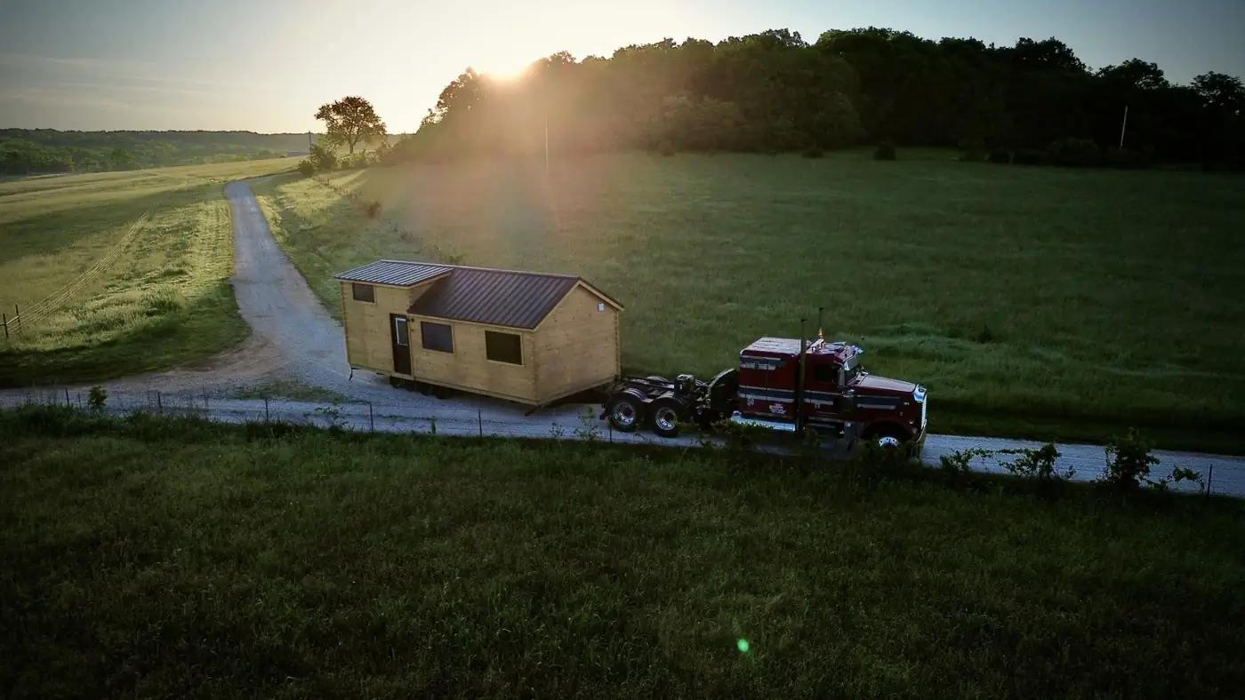 Red semi hauling a small cabin down a country lane past green fields at sunrise