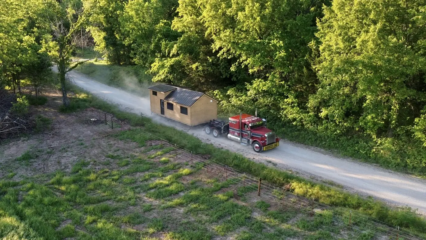 Aerial view of a red semi hauling a cabin down a gravel road beside a line of trees