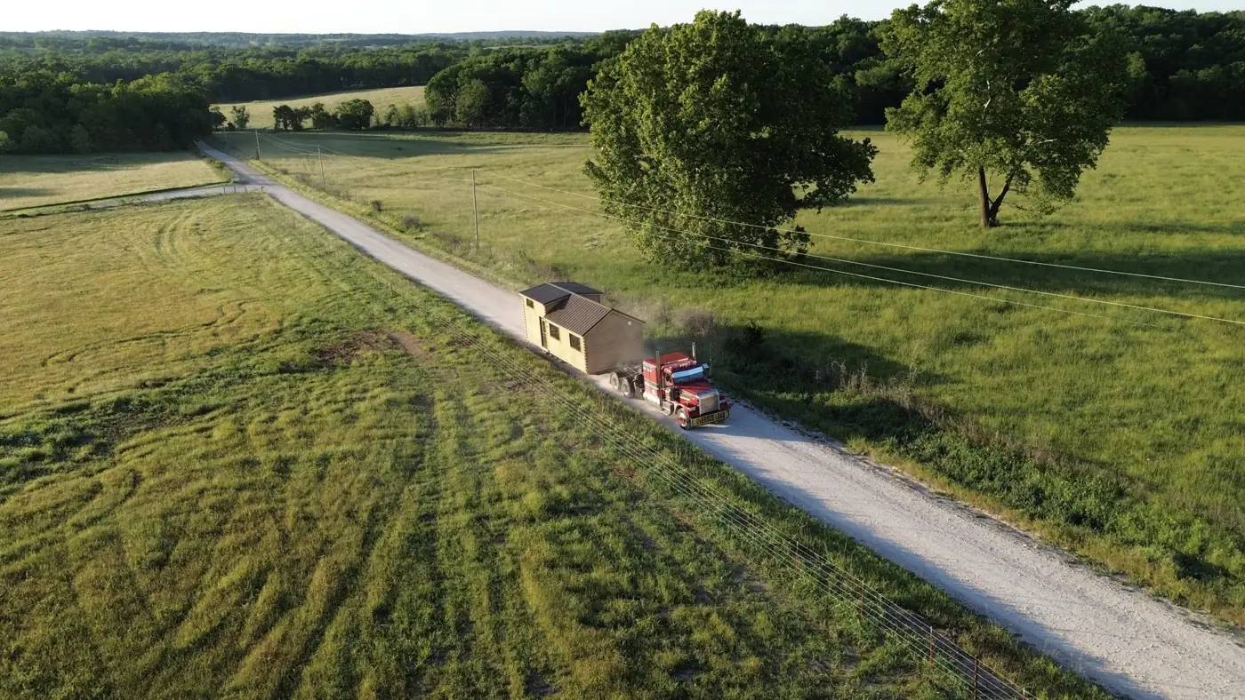 Aerial view of a semi hauling a cabin along a gravel road through farmland