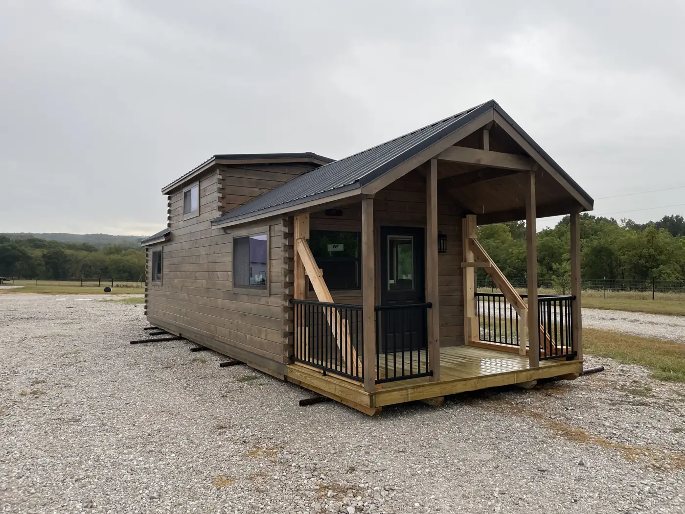 The Kodiak log cabin tiny home with a covered front porch, black railings and a loft, parked on gravel