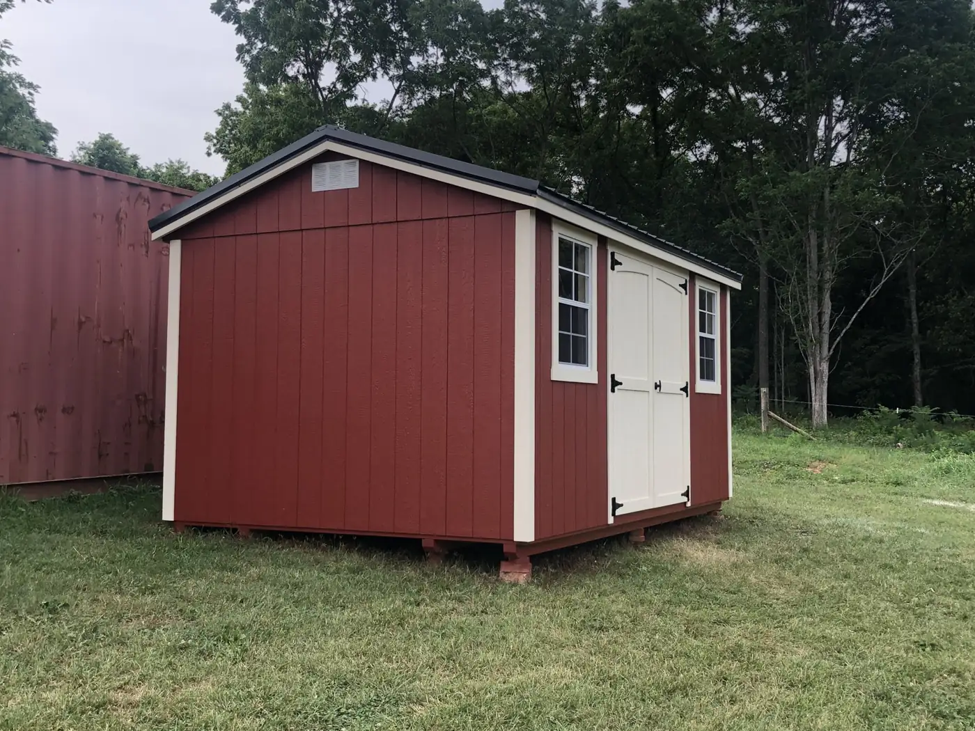 Barn red garden shed with white trim, white double doors and two windows on a grassy lot