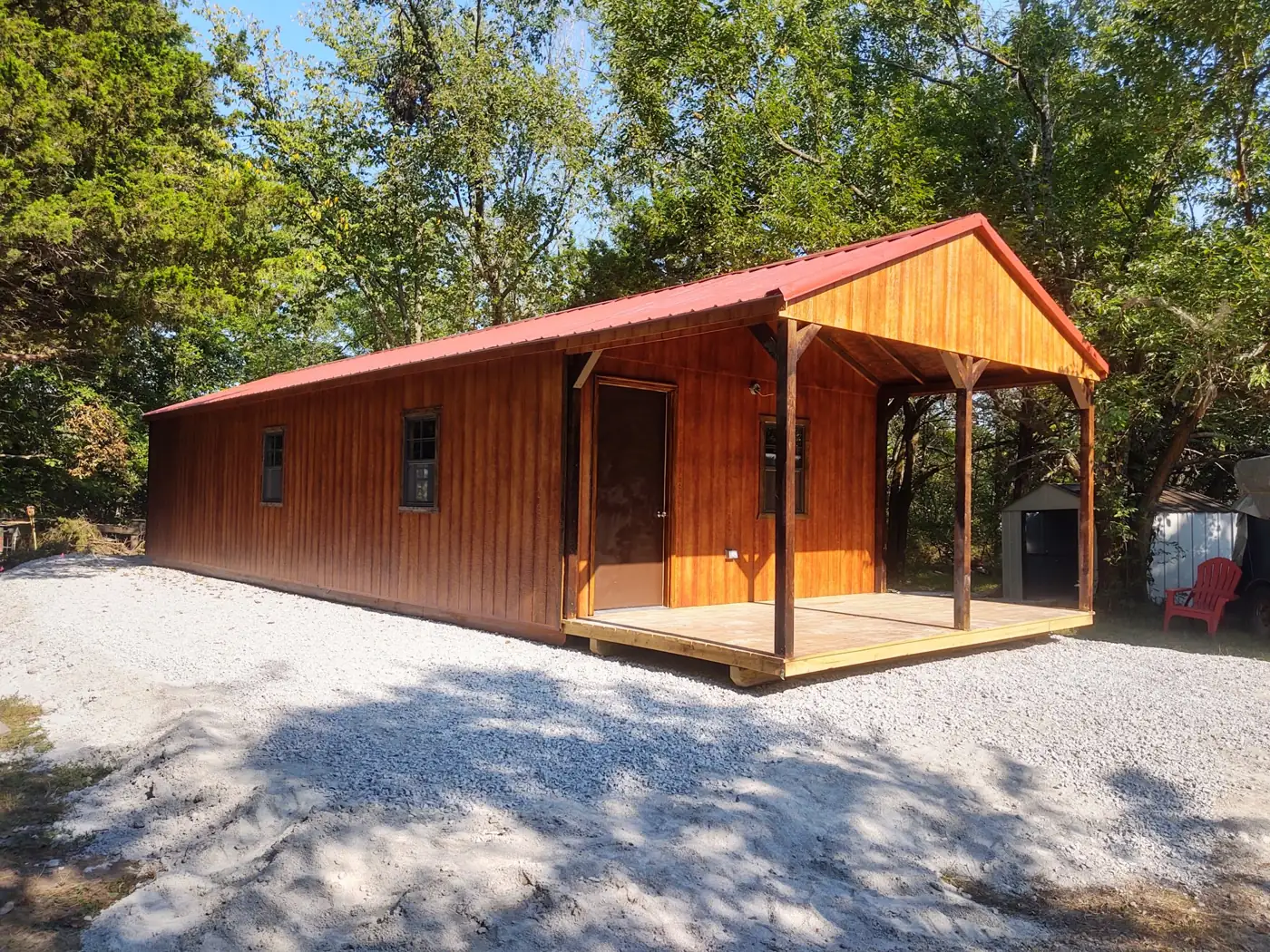 Long stained utility cabin with a red metal roof and covered porch on a gravel pad