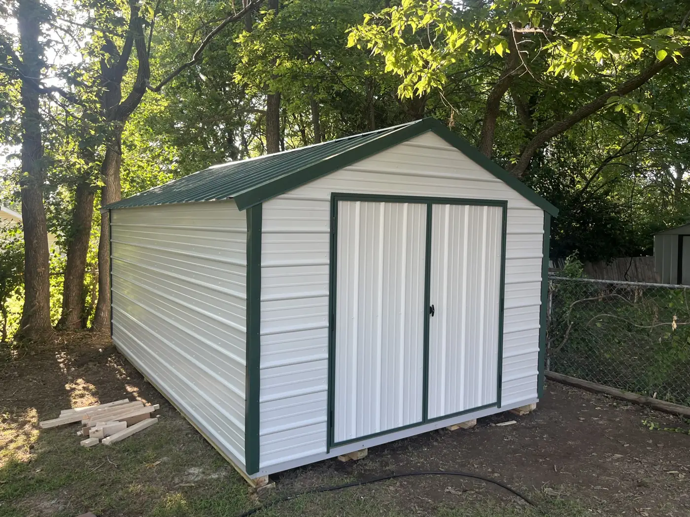 White utility shed with green trim and double doors set among trees